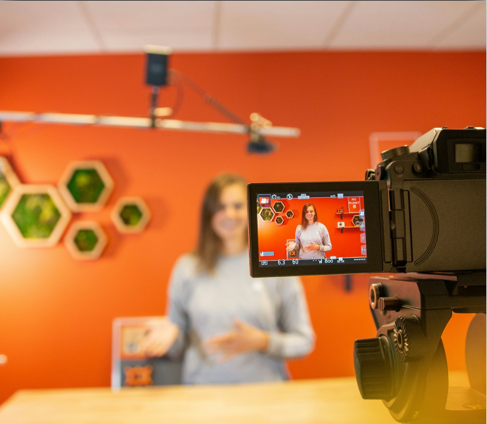 Camera recording a woman in a studio with a red background and hexagonal wall decor, capturing a corporate video for Edmonton.